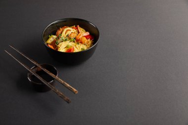 noodles with shrimps and vegetables in bowl near wooden chopsticks and soy sauce on black background clipart