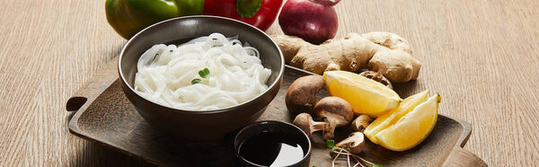 rice noodles in bowl near soy sauce, ginger root, onion and mushrooms on wooden tray, panoramic shot