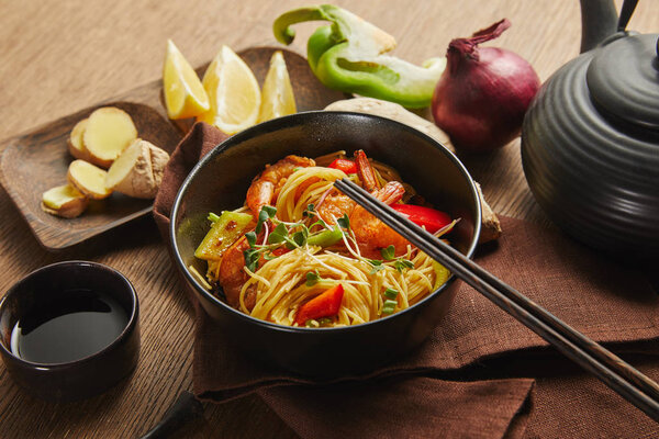 noodles with shrimps and vegetables in bowl near chopsticks, soy sauce, lemon and ginger root, teapot on napkin on wooden table
