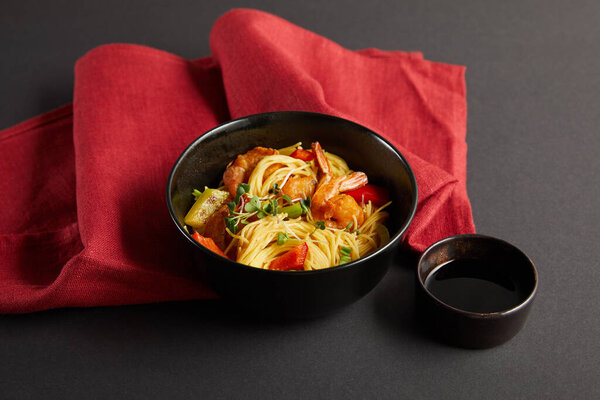 noodles with shrimps and vegetables in bowl near soy sauce on red napkin on black background
