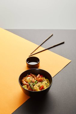 selective focus of noodles with shrimps and vegetables in bowl near chopsticks, soy sauce on black and yellow background isolated on grey clipart