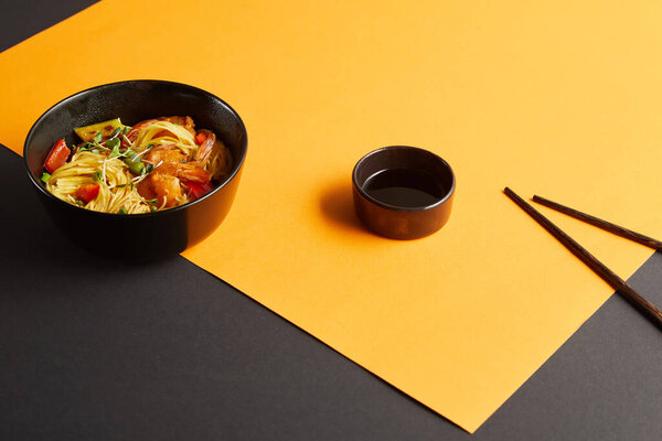 noodles with shrimps and vegetables in bowl near chopsticks, soy sauce on black and yellow background