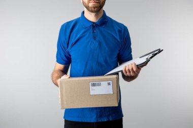 Cropped view of delivery man holding cardboard box and clipboard isolated on grey clipart