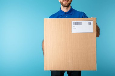 Cropped view of courier holding cardboard box with qr and barcode on card isolated on blue clipart