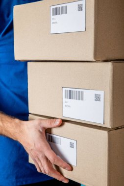 Cropped view of delivery man in uniform holding cardboard packages with qr codes and barcodes on cards clipart