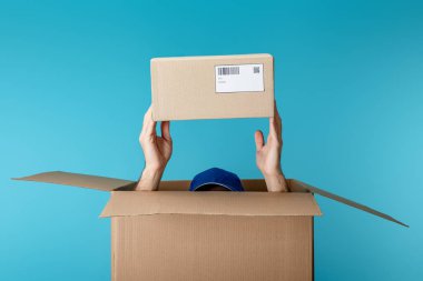 Cropped view of courier in cap holding cardboard package from box isolated on blue clipart