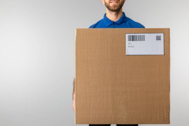 Cropped view of bearded courier holding cardboard package with qr code and barcode isolated on grey clipart