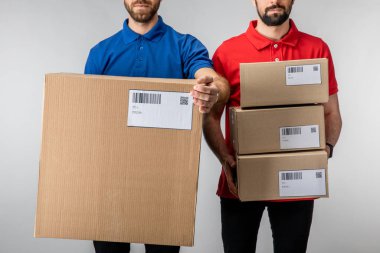 Cropped view of bearded delivery men holding boxes with barcodes and qr codes on cards isolated on grey clipart