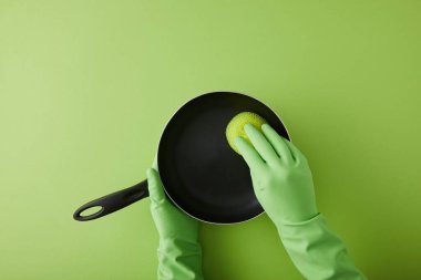 Lastik eldivenli temizlikçinin yeşil üzerinde süngerle tavayı yıkarkenki görüntüsü. 
