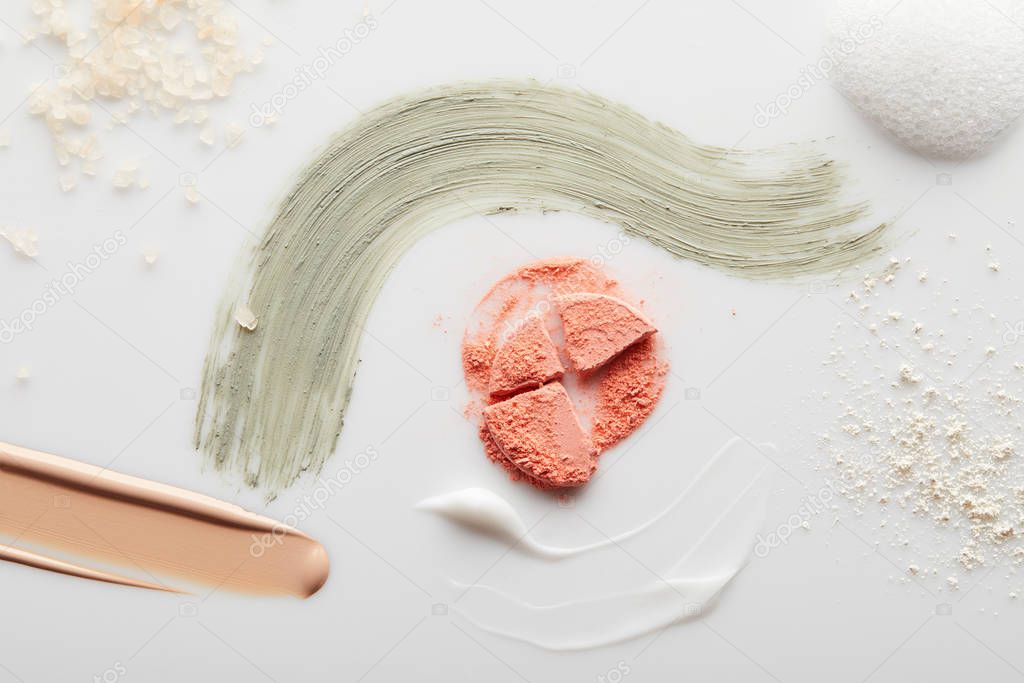 Top view of cosmetic, tone cream and clay brushstrokes with blusher, face powder, sea salt and soap foam on grey background