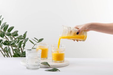 Yeşil bitkinin yakınındaki beyaz yüzeyde cam üzerine nefis sarı smoothie döken bir kadın manzarası.