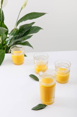 Yeşil barış çiçeğinin yakınındaki beyaz yüzeyde taze, lezzetli sarı smoothie bardakları gri üzerine izole edilmiş.