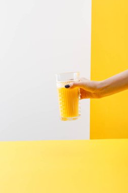 Gri ve sarı arka planda taze, lezzetli sarı smoothie bardağı tutan bir kadın görüntüsü.