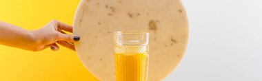 Gri ve sarı arka planda taze sarı smoothie, panoramik çekim, cam kenarında mermer daire tutan bir kadın görüntüsü.