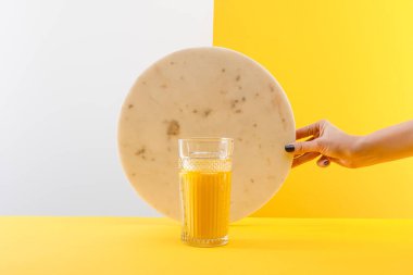Gri ve sarı arka planda taze sarı smoothie 'nin camının yanında mermer daire tutan bir kadın.