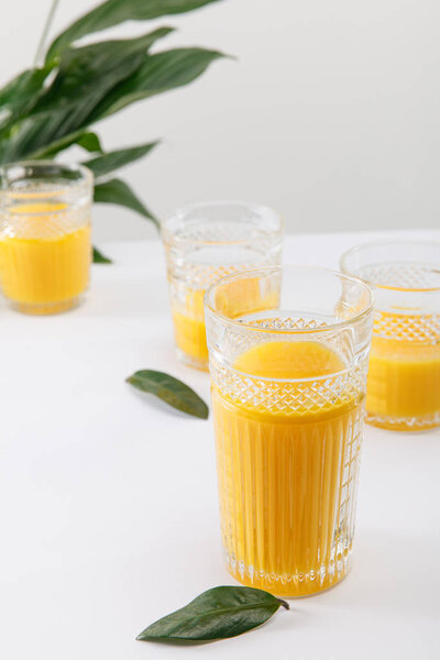 selective focus of glasses of fresh delicious yellow smoothie on white surface near green peace lily plant isolated on grey