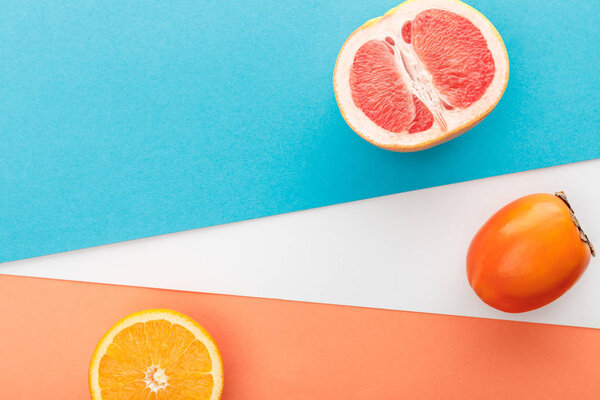 Top view of grapefruit half, slice of citrus fruit and persimmon on blue, orange and white background