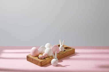 Ahşap tahtada paskalya yumurtaları, pembe ve gri arka planda dekoratif tavşanlar.