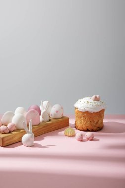 Paskalya yumurtaları ahşap tahtada, paskalya pastasının yanında dekoratif tavşanlar pembe ve gri arka planda.