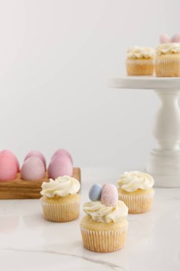 Delicious Easter cupcakes on surface and cake stand near painted chicken eggs on grey background  clipart