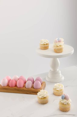 Delicious Easter cupcakes on surface and cake stand with painted chicken eggs on grey background  clipart