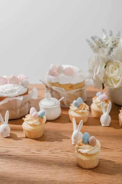 Selective focus of cupcakes with decorative rabbits, sugar bowl, easter cakes and vase with bouquet isolated on grey