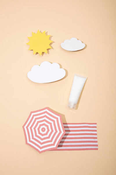 Top view of paper cut sun, clouds, beach umbrella and blanket with tube of sunscreen on beige