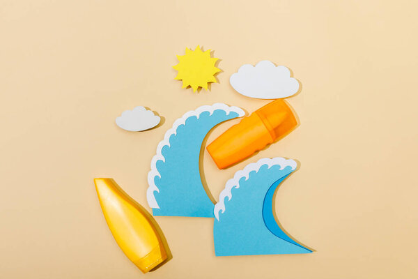 Top view of paper cut sun, clouds, sea waves with tube and bottle of sunscreen on beige 