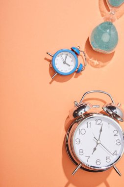 top view of alarm clocks and hourglass on peach background clipart