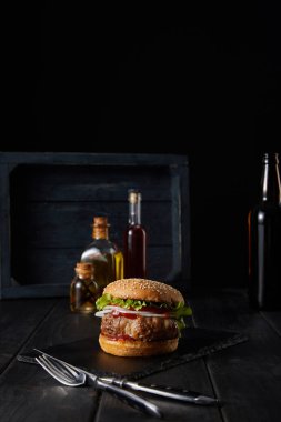 Hamburgerin siyah kesim tahtasına odaklanması siyah çatal bıçak, yağ, sirke ve bira şişelerinin yakınındadır. 