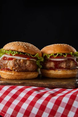 İki hamburger ahşap doğrama tahtasının üzerinde kareli masa örtüsünün üzerinde, siyah üzerine izole edilmiş.