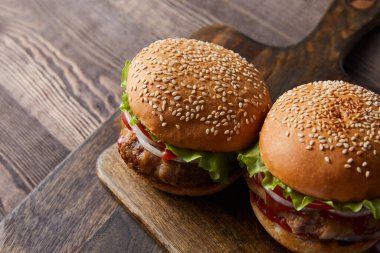 Ahşap kesim tahtasındaki iki burgerin yüksek açılı görüntüsü, çatal bıçak, yağ, sirke ve bira şişeleri siyah üzerine izole edilmiş. 