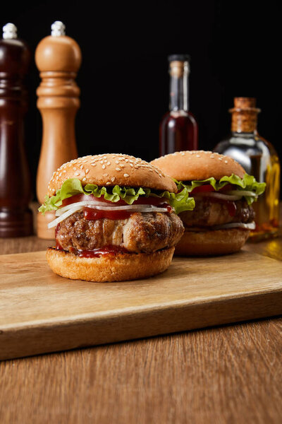 selective focus of two burgers on wooden cutting board with oil, vinegar and beer bottles, pepper and salt mills isolated on black