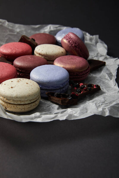 close up view of assorted delicious colorful french macaroons on crumpled paper with chocolate on black background