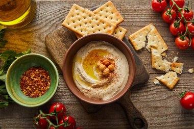 Top view of crackers, bowls with hummus and spices, vegetables, parsley and jar of olive oil on wooden background clipart