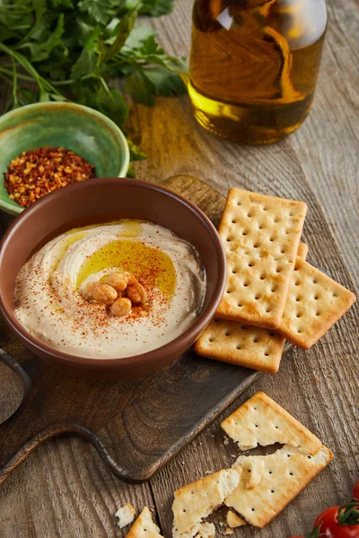 Selective focus of crackers, parsley, bowls with paprika and hummus on cutting board on wooden background