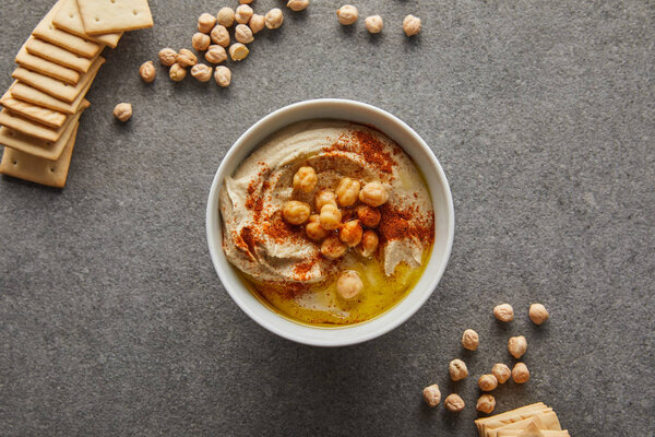 Top view of bowl with delicious hummus, chickpea and crackers on grey background