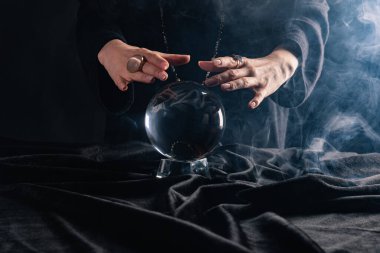 Cropped view of witch performing ritual with crystal ball on black background clipart