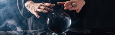 Cropped view of witch performing ritual with crystal ball on black, panoramic shot clipart