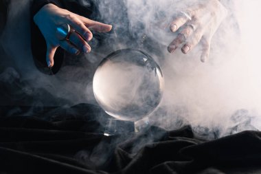 Cropped view of female hands above crystal ball with smoke on dark clipart