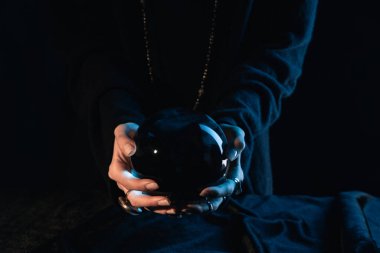 Cropped view of witch with outstretched hands holding crystal ball on black background clipart