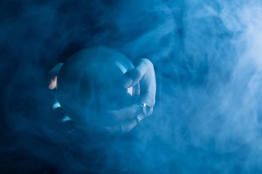 Cropped view of female hands with crystal ball and smoke around on dark blue background clipart