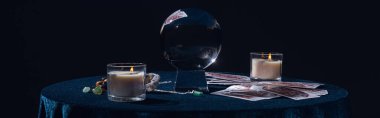 KYIV, UKRAINE - JANUARY 9, 2020: crystal ball with candles, tarot cards, stones and chain on round table isolated on black, panoramic shot clipart