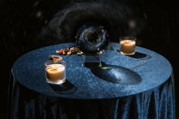 Crystal ball near candles and fortune telling stones on dark blue velour cloth on black