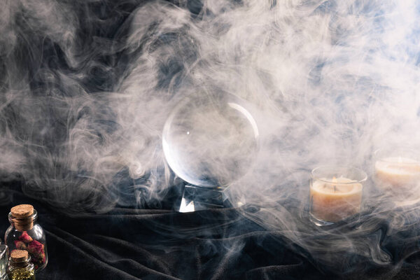 Crystal ball, candles, jars of herbs and buds with smoke around on dark background