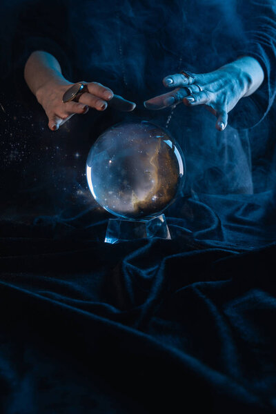 Обрезанный вид ведьмы, выполняющей ритуал с хрустальным шаром на темно-синем
