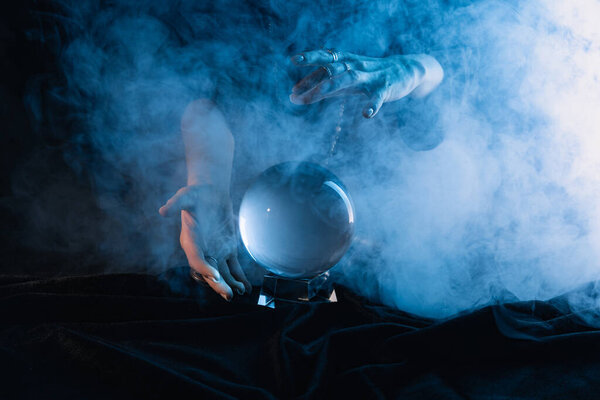 Обрезанный вид ведьмы, выполняющей ритуал с хрустальным шаром на темно-синем фоне
