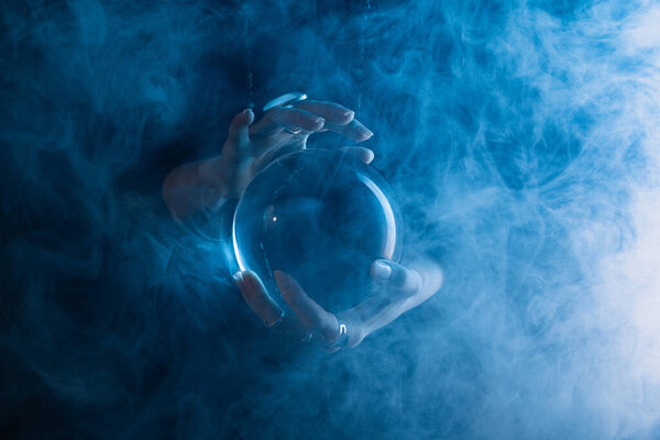 Partial view of witch holding crystal ball with smoke around on dark blue 
