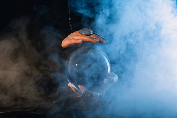 Partial view of female hands with crystal ball and smoke around on dark blue 