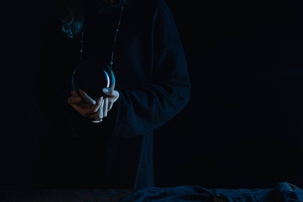 Cropped view of witch with clenched hands holding crystal ball on black background